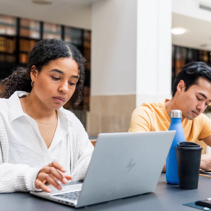 Two people sit at the public desks in the British Library, one working on their laptop, the other writing on a notepad. 