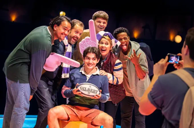 The cast of The Little Big Things gather around Henry who holds a rugby ball, they pose for a photo