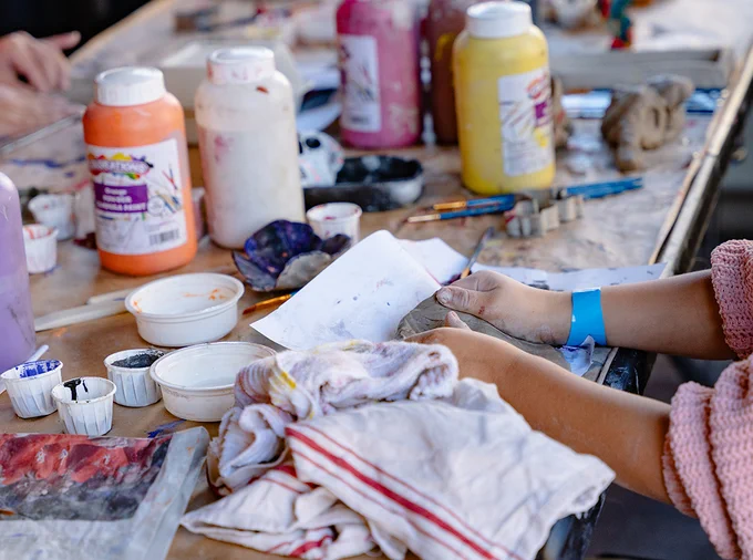 Close-up photograph of a child's hands as they mold clay on a table covered in paints and art supplies.