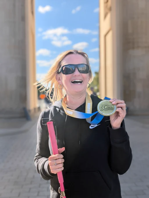 The image shows a woman standing outdoors, smiling broadly. She is wearing sunglasses and a black Nike hoodie. She is holding a pink cane in her left hand, which suggests she is visually impaired. In her right hand, she is holding a medal with a blue and yellow ribbon around her neck. The medal has the text "50 BMW BERLIN-MARATHON" on it. The background features a clear blue sky with some clouds and a large stone structure, possibly a monument or an archway. The woman appears to be celebrating an achievement, likely completing the Berlin Marathon.