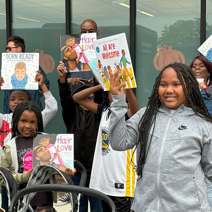 A group of children and adults pose with smiles holding the books they received from the Banned Wagon. 