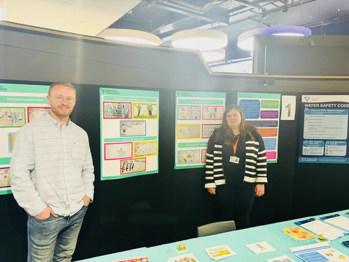 James is on the left wearing a white shirt and denim jeans. Paula is on the right wearing a stripy black and white jumper and black jeans. Behind James and Paula there are 3 posters showing children's drawings. There is also a Water Safety Code poster from Water Safety Scotland. 