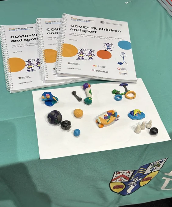 Three copies of the covid-19, children and sport report are sitting on a table. Below the report are clay models made by children. The models depict a swimmer, weight bars, bicycle, balls, skateboard, bowling pins and bowling ball. 