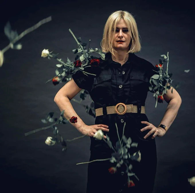 A headshot of Claire Welles wearing a black short-sleeved shirt dress with brown belt around the waist, and hands on her hips. The background is black and red and white roses fall in front of her.