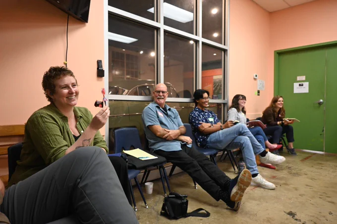 Five people sit in small blue plastic chairs, all laughing and smiling. The person closest to the camera holds up a small, lollipop-like device.