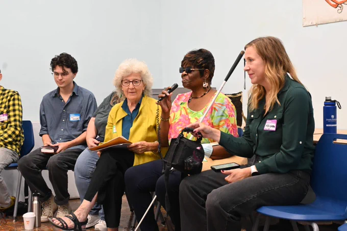 Five people sit on blue plastic chairs. A Black woman wearing sunglasses holds a potable microphone in one hand and a guide stick in the other while everyone else listens. 