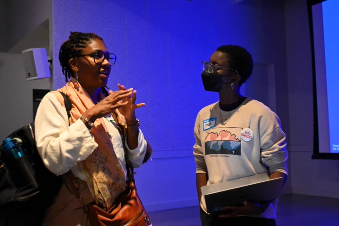 Two women talk to each other in a blue-lit room. The woman on the right wears a mask and a pin that says “Afro Charities”. 