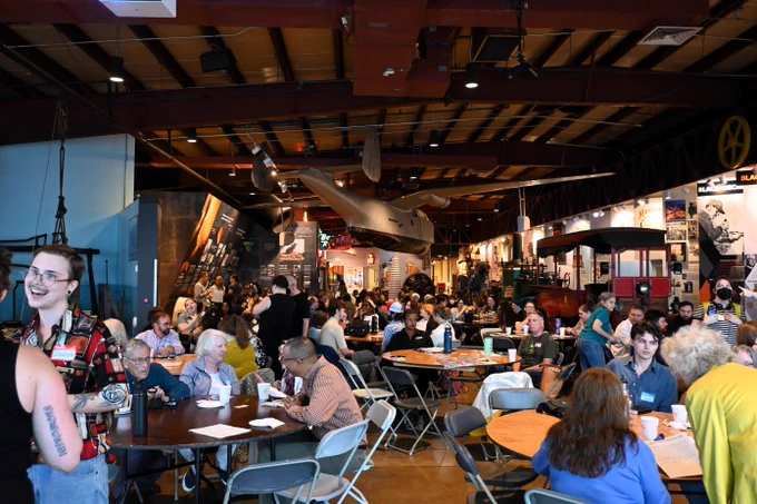 A large room filled with people talking at a dozen large round tables and walking around the room. The room’s walls and ceiling hold exhibits displaying large-scale models of an airplane and a Baltimore streetcar, amongst other industrial-related artifacts. 