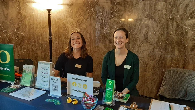 UO Libraries' employees staff a promotional table in Knight Library. The booth features informational brochures, a sign saying "FOLLOW US ON @UOLIBRARIES," a bowl of candies, and other prizes.