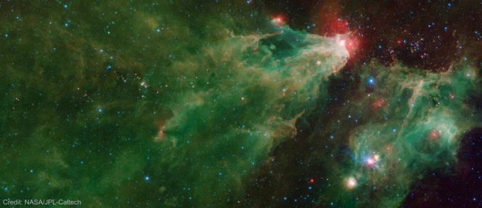 This image was compiled using infrared data from NASA's Spitzer Space Telescope. Most of the image is filled with a triangular wispy green-and-orange shape, which is a nebula, or a cloud of gas and dust. A bright region at the tip of the nebula glows brightly because it has been heated by the stars' radiation, which creates a surrounding red glow. On the left side of this image, a dark filament runs horizontally through the green cloud. A smattering of baby stars appear inside it as red and yellow dots. A smaller glowing green nebula on the right side of the image includes a blue star crowned by a small, red arc of light. The image is watermarked with the words “Credit: NASA/JPL-Caltech.”