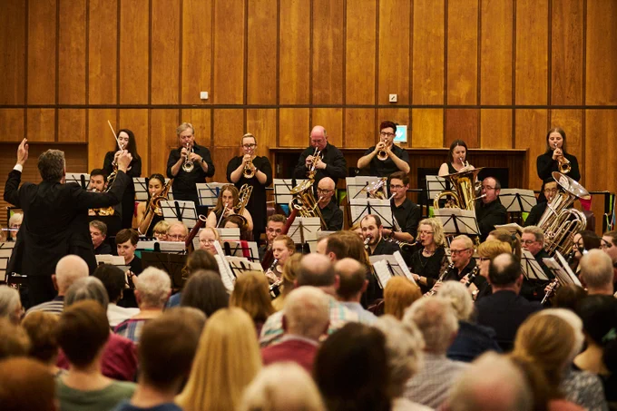 Photo of brass band playing for an audience. The players are playing an array of brass instruments with sheet music in front of them. There a conductor with arms raised, mid-conducting. The audience can be seen in the foreground of the photo. The room has a warm ambience and the walls have wood panelling. 