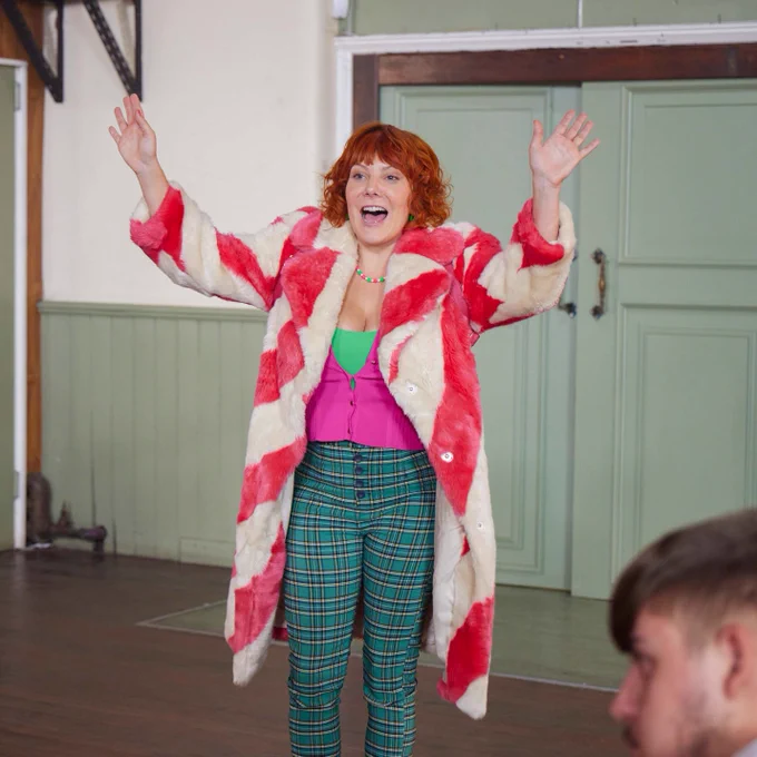 Alma (Sophie Willan) walks into a room with her hands held in the air, mid-speech. Alma wears a green top and trousers and a furry striped pink and white coat.