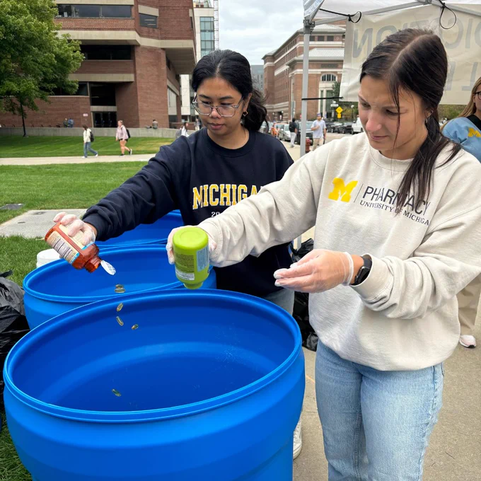 Two students dumping unused pills into a blue container outdoors. 