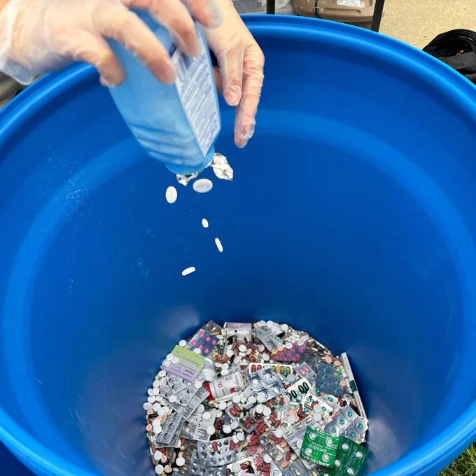 A person pouring a bottle of pills into a large blue container filled with various types of medication packages.