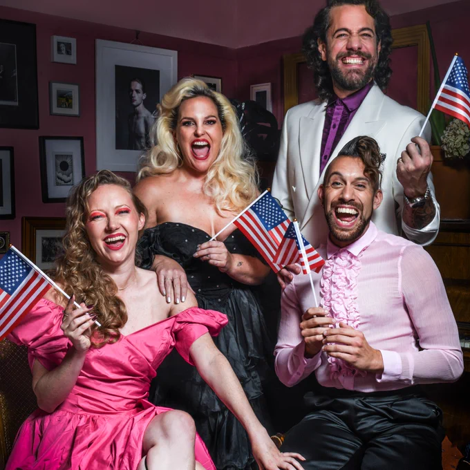 The four members of SpitLip (L-R: Tash, Zoe, Felix and David) done up all fancy and dressed in poofy gowns and frilly shirts as if going to a High School Prom They are all smiling excitedly and waving little US flags. It’s a vibe.