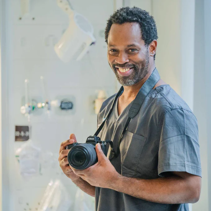 Karel is standing and smiling while having his profile taken. He is holding a camera in his hands that is strapped around his neck, while standing in a clinical setting and wearing scrubs. 