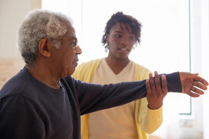 An older man receiving help with being physically active from a younger woman, who is supporting him to lift his arms.