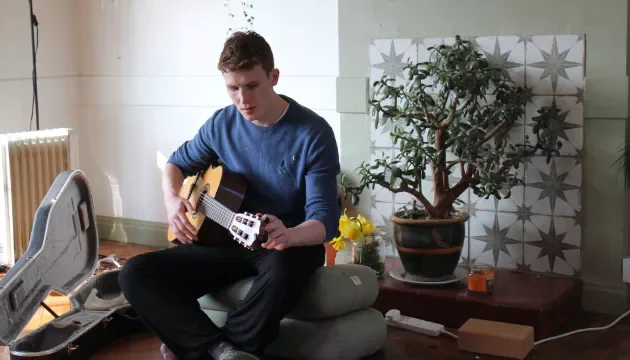Will Crawford sits on cushions tuning an acoustic guitar in a living room with a potted plant, a radiator, and a guitar case nearby.
