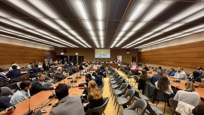 A photo of a conference room at the Palais des Nations in Geneva, Switzerland where an event commemorating the UN International Day of Older Persons is taking place. The room is packed with attendees.