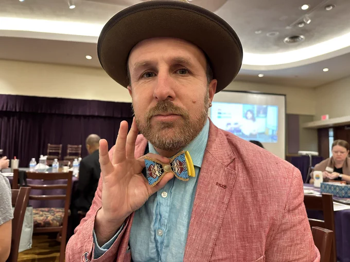Brian Redcorn in a cool peach jacket and blue shirt holding up a bowtie beat it by Lu Whitebear