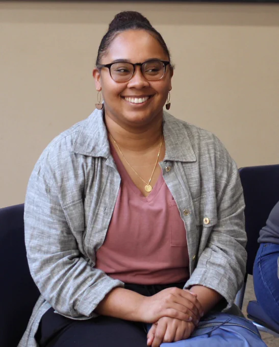 A smiling young black woman sitting down with her hands crossed on her lap. She is wearing glasses and looks happy.
