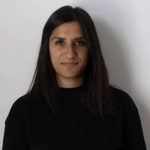 Reena is wearing a black top tucked into black trousers with shoulder length black hair. 
