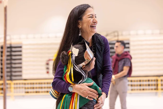 Secretary Haaland and a young Indigenous child smile while hugging. The Secretary wears a dark purple shirt, and the child's hair is decorated with a silver crown and red, green, yellow, and silver ribbons. 