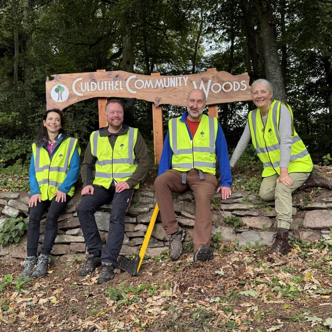 Four members of Culduthel Community Woods sit on a dry stone wall in front of a live edge timber sign which shows the name of their woodland and logo.