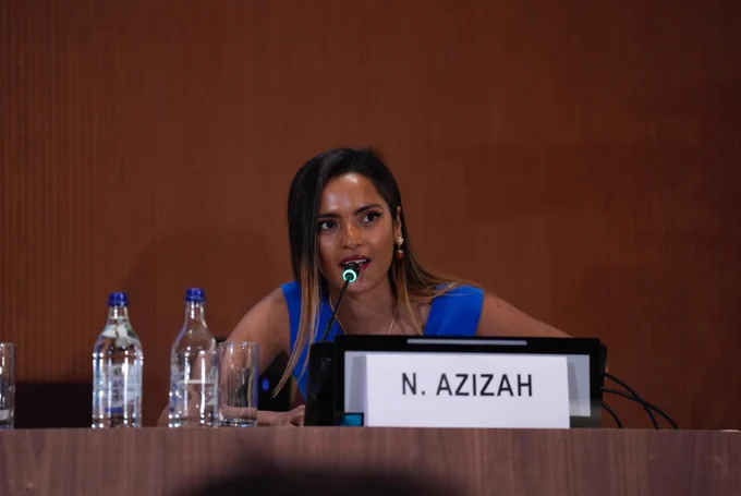 A woman in a blue sleeveless shirt sits on a podium as a speaker in front of a microphone, with the name card N. Azizah. Two water bottles are standing in front of her.