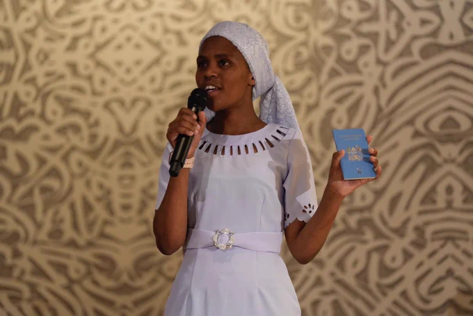 A woman wearing a white dress and head coverage is standing on what appears to be a stage, speaking into a microphone and holding a blue passport with the other hand. 