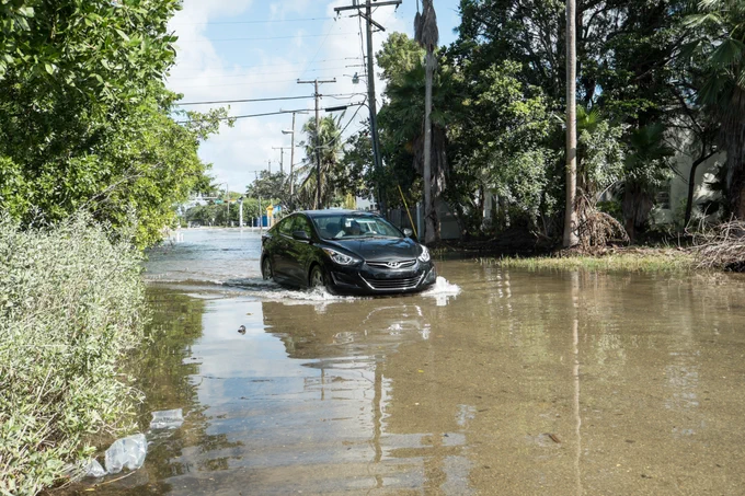 A street that is flooded with a black car going through it and trees along the side of the street.