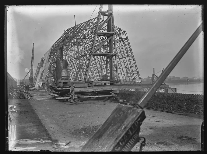Sankey photo 874 Airship Shed Under Construction 1910
Cavendish Dock looking East. A large steel structure is under construction alongside the dock wall. This is the Airship shed which will be used for the construction of the airship HMA No. 1, known as Mayfly. Steam powered winch on quayside, large crane behind shed. Various industrial buildings and tall chimney probably the Vulcan Steel and Iron Works in the background.
