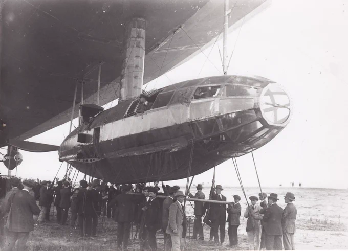 Sankey photo 1906 Vickers Airship, R80, Walney Island. The Forward Gondola 1920
The forward gondola of the airship with an inflated landing buffer underneath. One of the rear wing cars in view in the background. Many men holding guy ropes. Men in the gondola and a portion of the propellor in view. 
