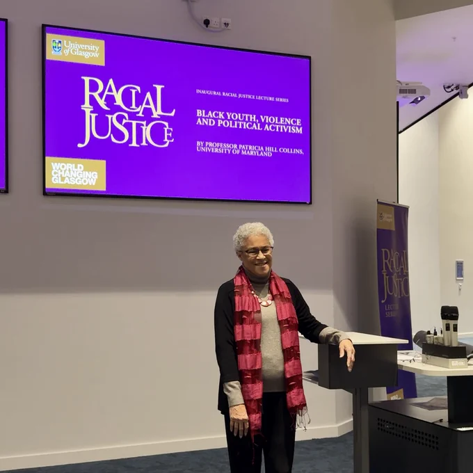 Patricia Hill Collins stands for a photograph with a purple, white and gold graphic in the background on a screen that reads: Inuguaral Racial Justice Lecture Series Racial Justice Black Youth, Violence and Political Activism by Professor Patricia Hill Collins, University of Maryland 