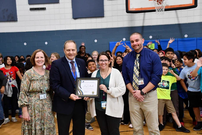 Sycamore Park Elementary School Assistant Principal Amy Cackowski stands with Superintendent Tony Brads and two other staff members during an award presentation. Cackowski is holding a framed certificate for the CCPS Spotlight Award, recognizing her contribution in organizing the division's Back to School Bash. A group of students is gathered behind them in a gymnasium, smiling and waving at the camera.