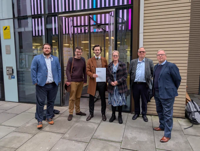 6 people from different AHP bodies standing outside the Department for Health and Social Care. The person in the middle holds a letter.