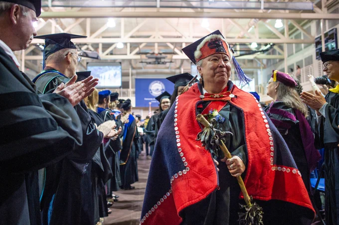 A person in traditional Indigenous regalia, including a feathered headdress and red shawl, walks proudly at a graduation ceremony, receiving applause from faculty members in academic robes.