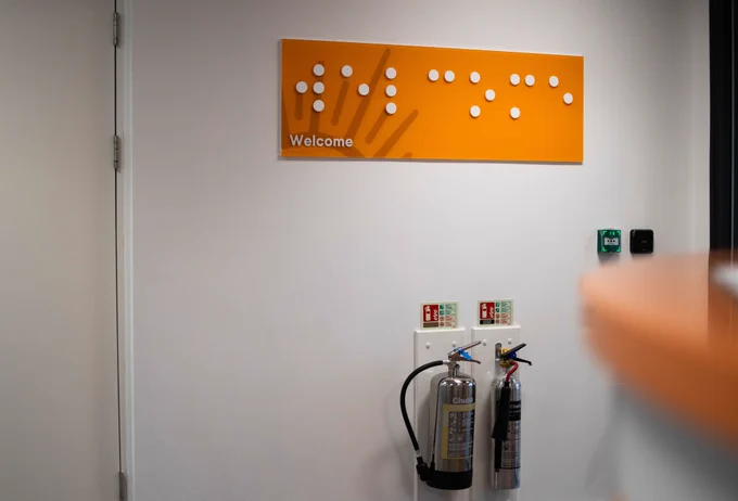 A white wall with a large orange sign on the wall which has white, braille on it and the word welcome written in english underneath.