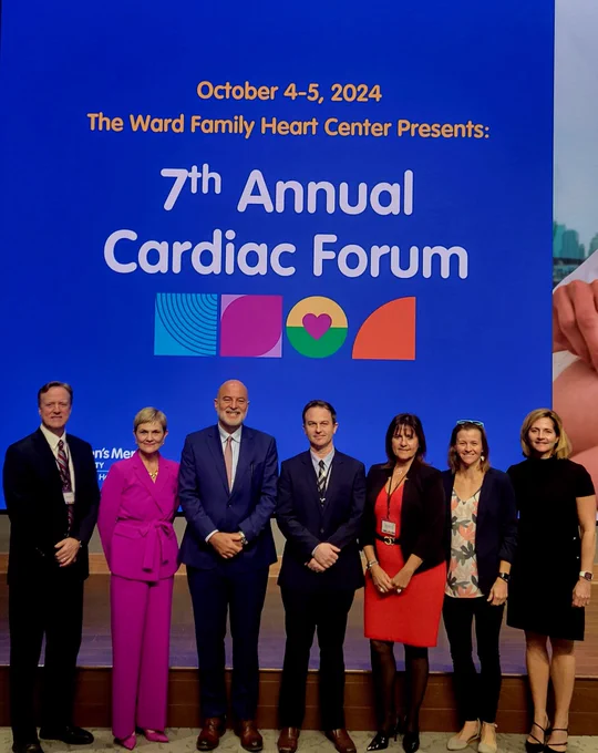 Group of seven individuals standing in front of a banner for The Ward Family Heart Center's 7th Annual Cardiac Forum, dated October 4-5, 2014.