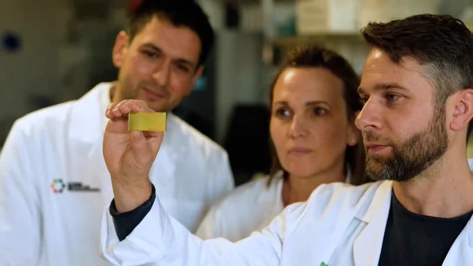 L - R: AlleSense co-founders Prof Brian Abbey, Prof Belinda Parker and Dr Eugeniu Balaur looking at a NanoMSlide. They are standing inside a lab, wearing lab coats.