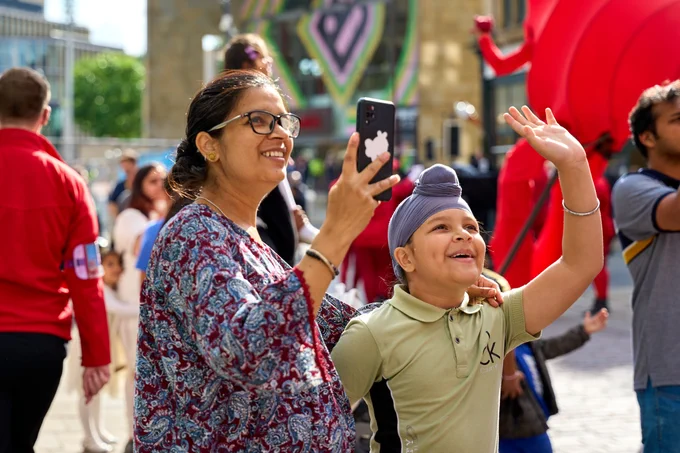 Two individuals, one with a smartphone, smiling and gesturing excitedly. In the background, there is a crowd and a large red giraffe puppet. 