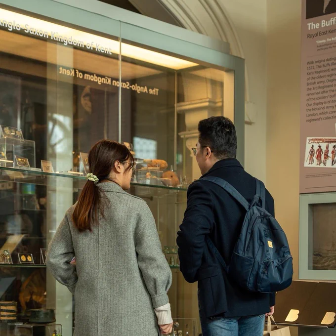 Two people standing in front of a museum display case filled with artefacts. One person looks ahead at the objects whilst the person they are with is turned towards them as they talk about the items on display.