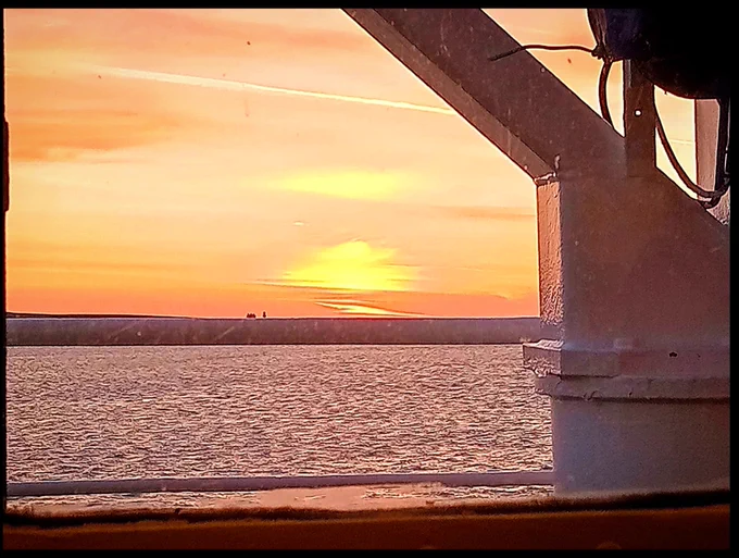 The sunrise in Orkney this morning. The sun rising over a strip of land on the horizon. The sky is orange. In the foreground is the deck and structure of the ferry.
