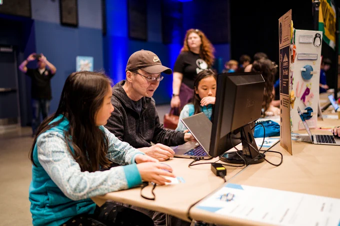 A mentor behind a screen with a young coder sat on each side of them.