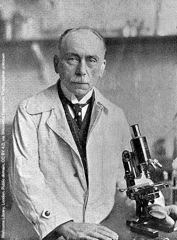 Black and white photo of Jules Bordet looking directly at the camera. He is in a laboratory wearing a lab coat and sitting in front of a microscope.