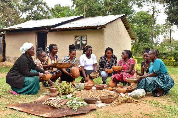 A group of women sat in a semi-circle, holding up pots with crops and seeds. 