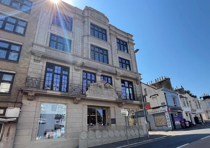 Image of the exterior and entrance of the Observer Building in Hastings. Blue sky in the background and image of buildings and shops on the right