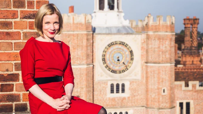 A photo of Lucy Worsley wearing a red dress and red lipstick, sitting against a red brick wall on the roof of Hampton Court Palace with the astronomical clock in the background