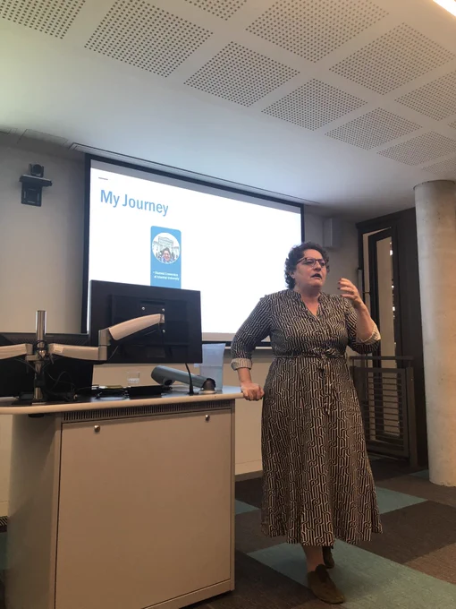 Woman in striped dress speaks in front of screen with slide reading ‘My Journey’