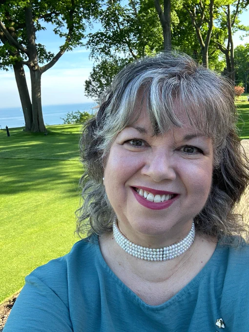 A white woman with shoulder-length grey hair smiles on a beautiful sunny day. She is wearing a pearl choker and a blue top.